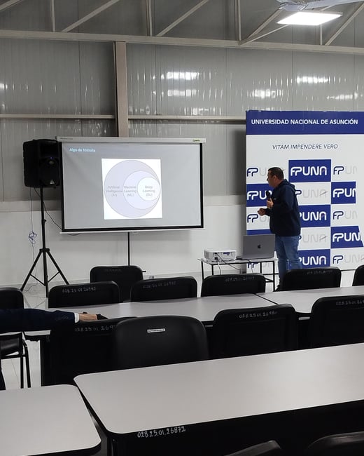 A man stands in a classroom next to a projector screen presenting a diagram on Artificial Intelligence, Machine Learning, and Deep Learning, with a banner for the Universidad Nacional de Asunción in the background.
