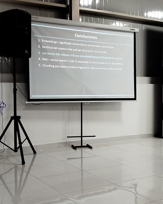 A photograph of a projector screen displaying a Spanish presentation slide titled 'Conclusiones' inside a room with a tiled floor, alongside a black speaker on a tripod stand.
