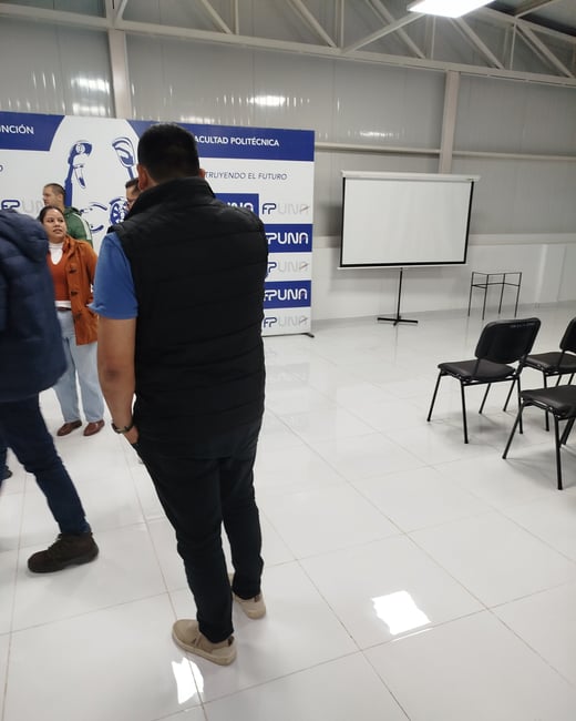 An indoor shot of a presentation hall or classroom featuring a white tiled floor, a projection screen, chairs, and a banner for 'FACULTAD POLITÉCNICA FPUNA' with several people standing and talking.