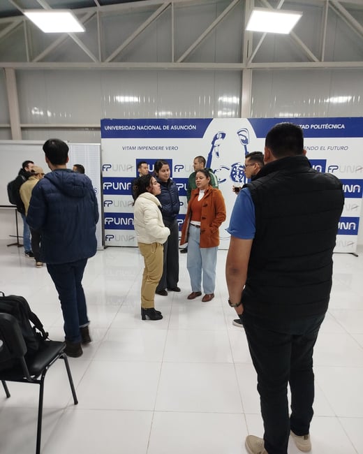 A group of people standing and conversing in a brightly lit indoor hall at a university event, in front of a blue and white FPUNA backdrop.