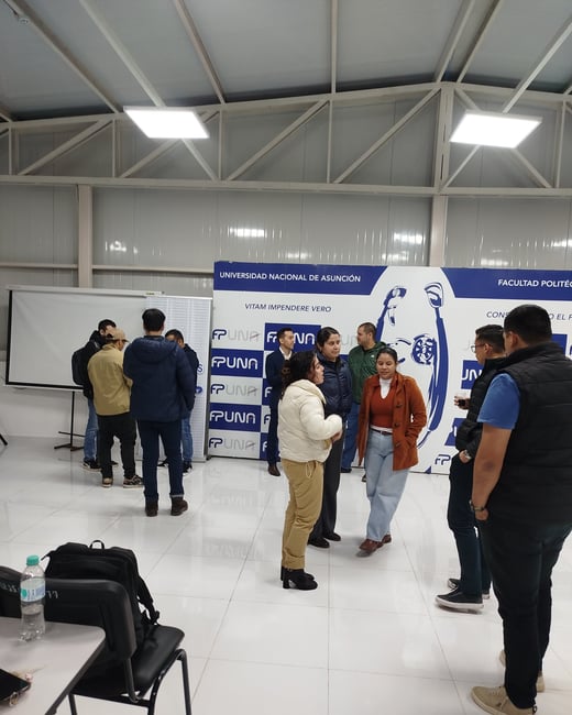 A group of people are gathered in an indoor lobby or hall, socializing and discussing near a large blue and white banner for the Universidad Nacional de Asunción.