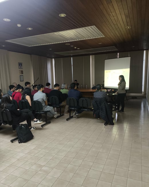 A photograph captures an indoor presentation or lecture where a woman stands next to a projection screen addressing an audience seated in rows of chairs. The room features a dark wood-paneled ceiling and light tiled floors.
