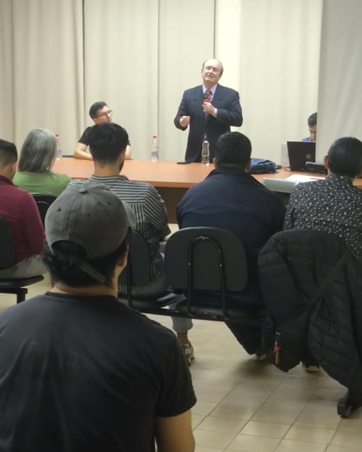 A candid photograph taken from the perspective of the audience shows a man in a dark suit giving a presentation with a microphone in a lecture hall or conference room.