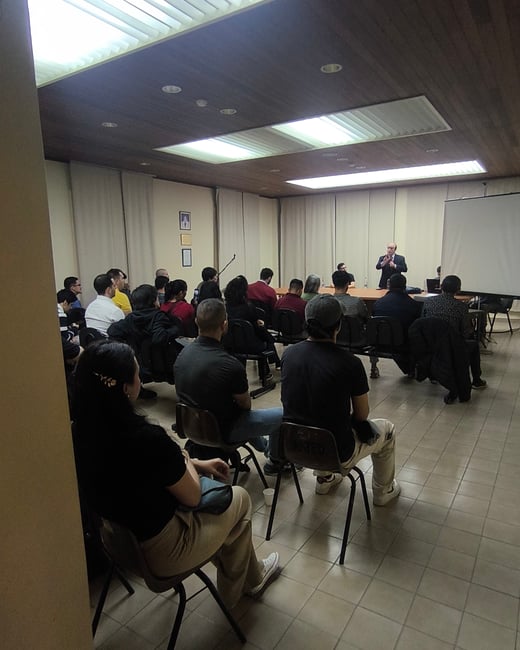 A candid photograph captures an audience seated in a dimly lit conference room, watching a man in a dark suit give a presentation near a blank projection screen.