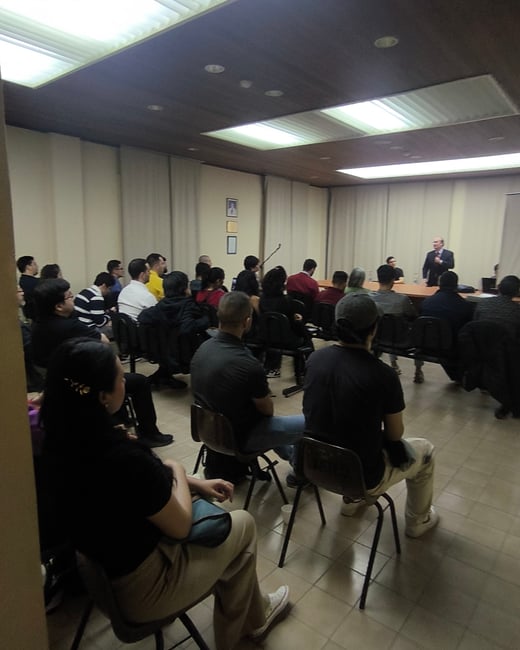 A candid photograph taken inside a dimly lit conference room or lecture hall, showing a large audience seated and facing a speaker who is standing at a table in the distance.