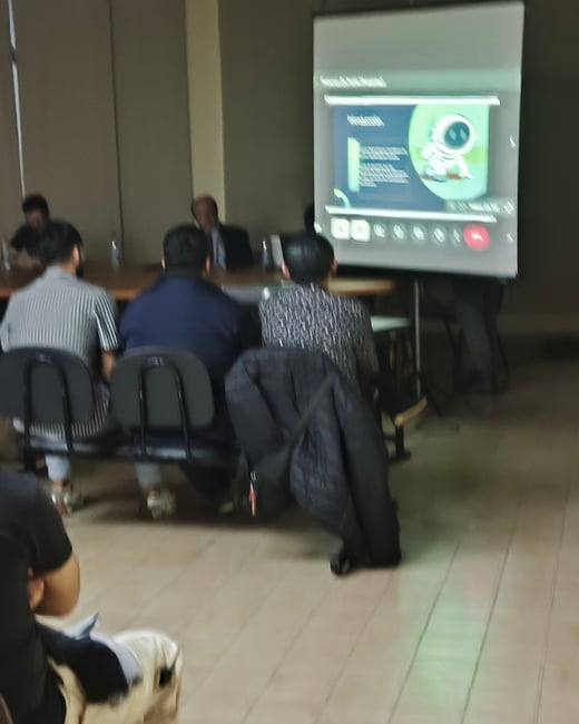 A blurry, low-light photograph of an indoor meeting or presentation, showing several attendees seated with their backs to the camera, facing a large projection screen.