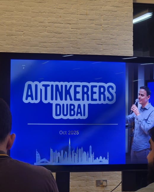 A photograph captures an indoor event setting where attendees are viewing a large screen displaying a presentation slide for the 'AI Tinkerers Dubai' event, scheduled for 'Oct 2025.' The screen shows a speaker on the right, and the backs of two audience members are visible in the foreground.