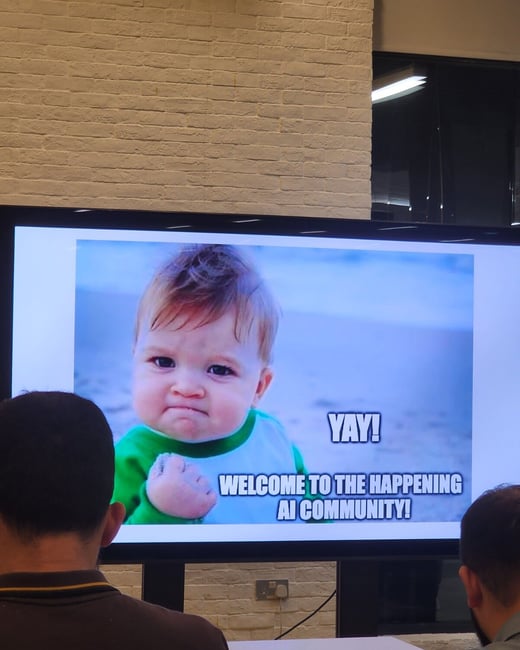 A photograph capturing an indoor presentation where a large screen displays the 'Success Kid' meme with text welcoming attendees to an AI community event. Two people are visible in the foreground, seen from behind, watching the screen.