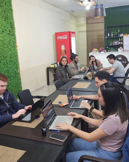 A group of people are seated at long tables in a cafe-like setting, all focused on their open laptops.