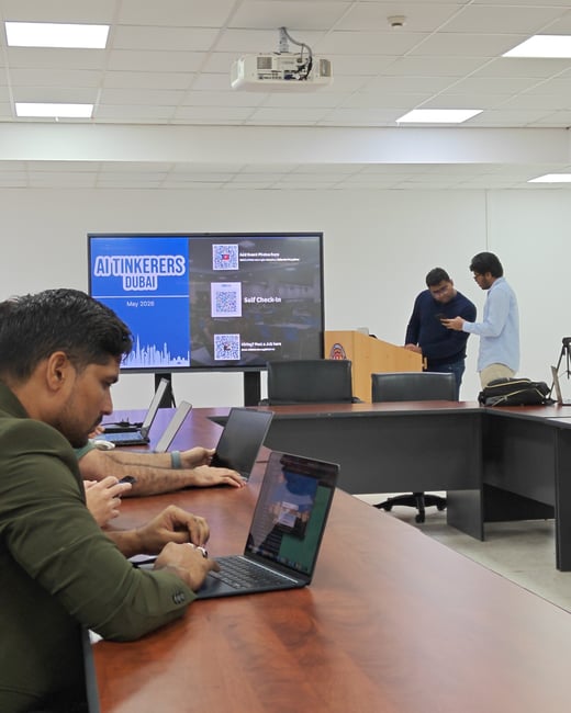 An indoor photograph of a conference room where several individuals are gathered, featuring a large screen displaying 'AI Tinkerers Dubai' with QR codes, and attendees working on laptops or talking.