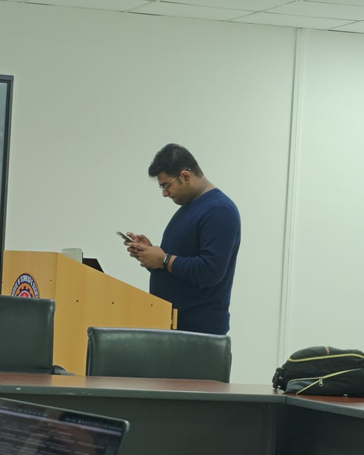 A man stands behind a wooden podium in a classroom, looking down at his smartphone with a laptop in the foreground.