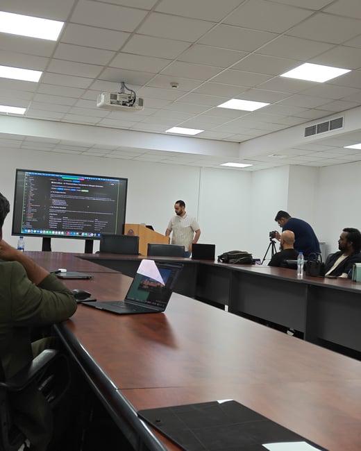 A presenter stands at a podium in a conference room, delivering a presentation shown on a large display screen to an audience seated around a wooden table.