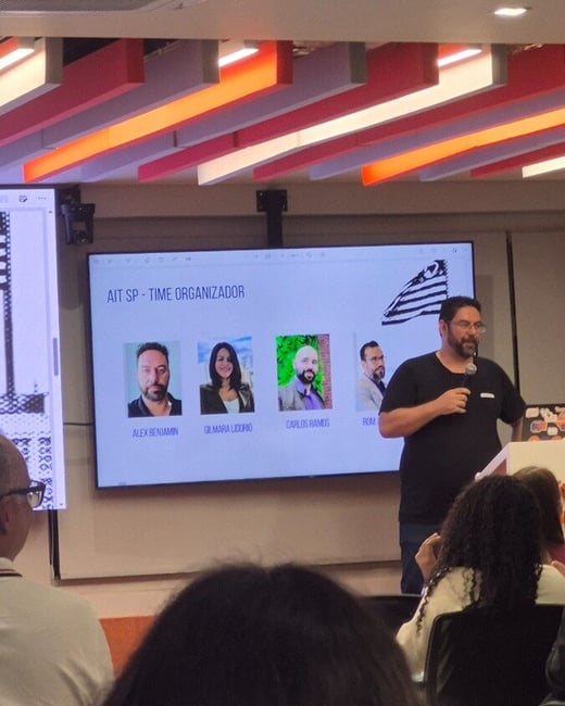 A man stands at a podium giving a presentation in front of a large screen displaying photos of team members.