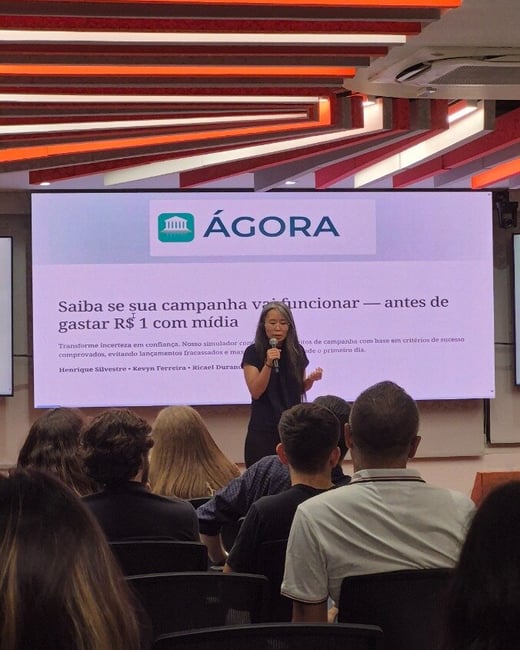 A woman is giving a presentation in front of a large screen displaying Portuguese text to an audience in an indoor setting.