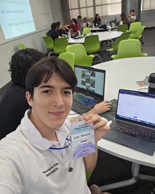 A young man takes a selfie while holding up his name badge at a 'Generative UI Global Hackathon' event, with other participants and laptops in the background.