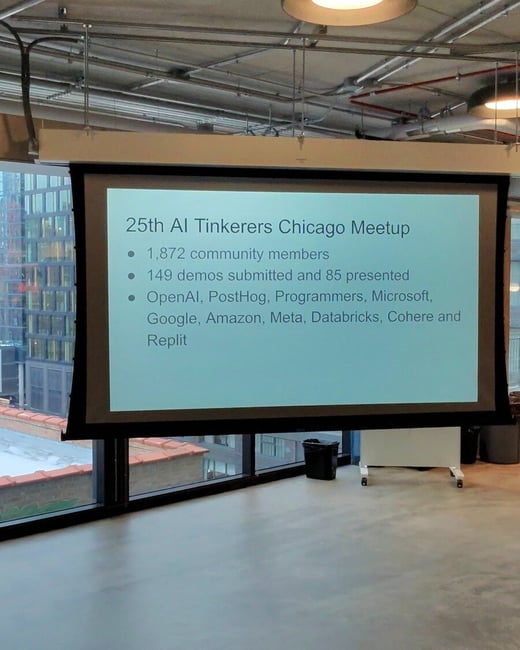 A projection screen in a modern office setting displays a slide for the 25th AI Tinkerers Chicago Meetup, highlighting community statistics and participating companies.