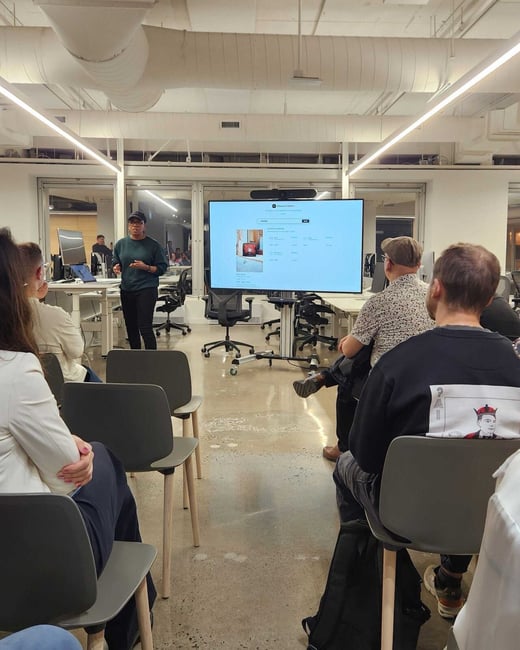 A photograph capturing a business presentation in a modern office space, featuring a presenter standing next to a large screen displaying a digital interface while addressing a seated audience.