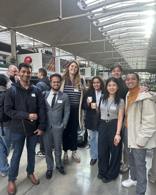 A group of seven young adults, featuring a mix of professional and casual attire, pose together for a photo inside a large, naturally lit event venue with a structured, industrial-style ceiling.
