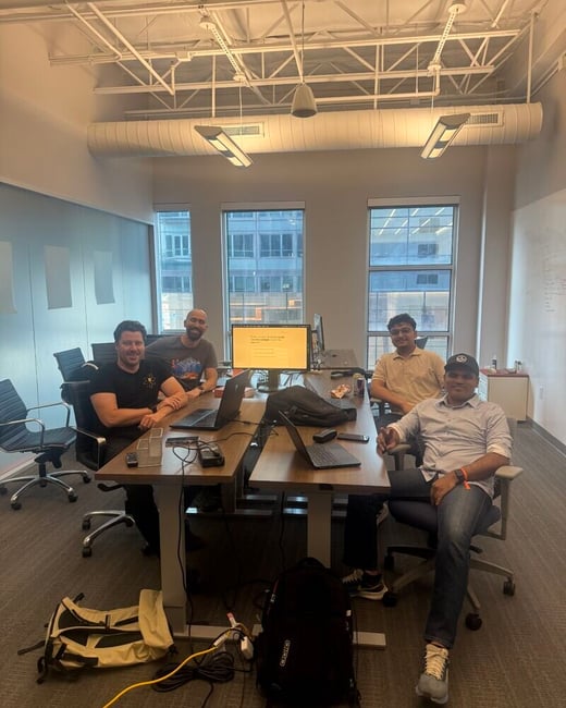 A group of four men are sitting at a desk in a modern office setting, posing for a photograph. They are surrounded by laptops, a monitor, and backpacks under an industrial-style ceiling.