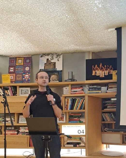 A man wearing a black shirt and dark jeans is giving a presentation or speech, standing behind a laptop stand and holding a microphone, in an indoor setting with a bright yellow floor and a wooden bookshelf backdrop.