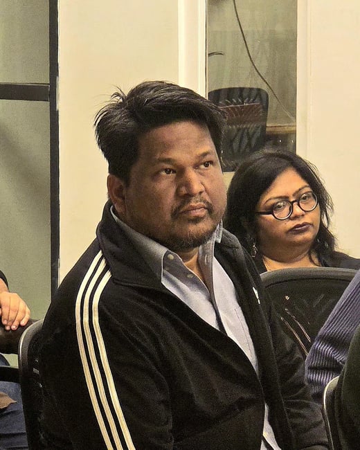 A close-up photograph of a man wearing a black zip-up jacket with three white stripes on the shoulder, seated indoors, with a woman wearing glasses visible behind him to the right.