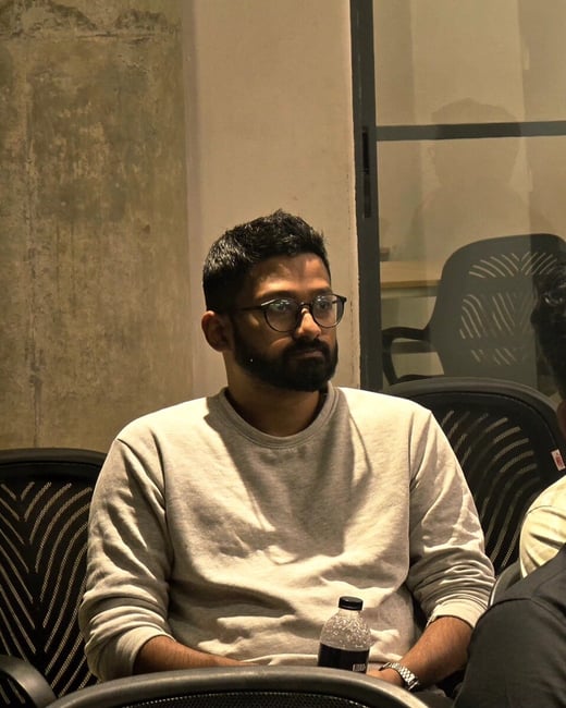 A candid indoor photograph of a bearded man wearing glasses and a light gray sweatshirt, seated in a chair and looking intently off to the left side.