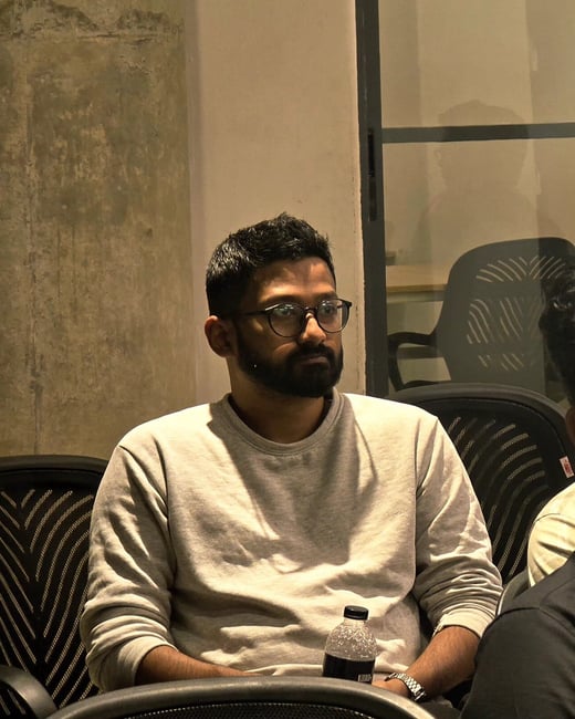 A candid, medium close-up photograph of a bearded man wearing glasses and a light gray sweatshirt, seated indoors in a low-light setting, looking intently off-camera.