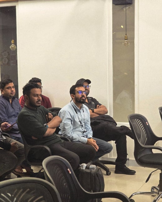 A candid photograph shows a group of men seated in black office chairs indoors, attending a meeting or presentation, with most of them having their arms crossed.