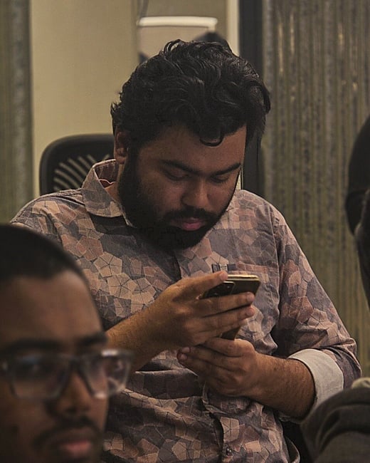 A close-up, candid photograph of a bearded man wearing a patterned shirt, sitting indoors and intently using a smartphone, with a blurred person visible in the immediate foreground.