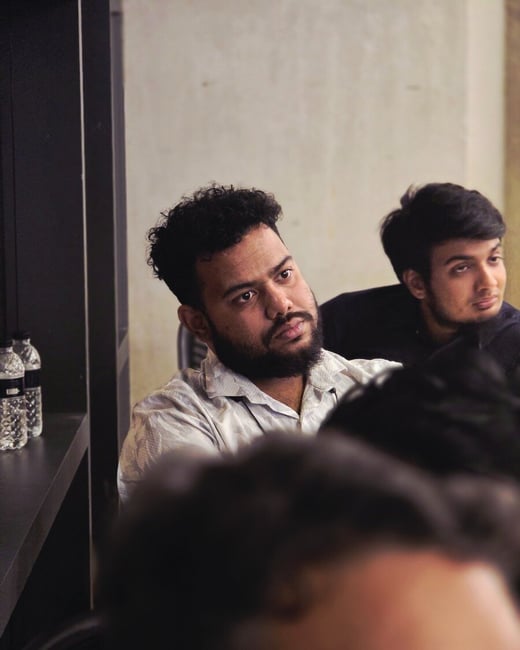 A candid indoor photograph capturing two men, focused on the bearded man in the foreground who is looking intently off-camera, while the back of a third person's head heavily blurs the bottom of the frame.