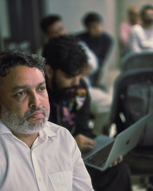 A candid, close-up photograph of a middle-aged man with a salt-and-pepper beard, wearing a white shirt, looking intently off-camera while seated in an audience; several blurred figures are visible behind him, one holding an open laptop.