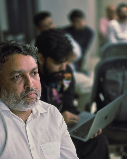 A close-up photograph of a middle-aged man with a gray beard wearing a white collared shirt, looking intently off-camera while attending an indoor event. Several other people, one holding a laptop, are visible but heavily blurred in the background.