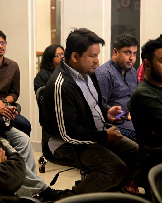 A candid indoor photograph showing several people, mostly men, seated in an audience setting, likely attending a meeting or presentation. The central figure wears a black zip-up jacket with white stripes and is looking down and to the side.