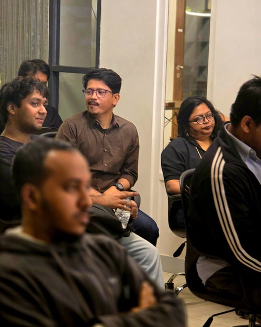 A candid photograph captures a group of people seated in an indoor setting, likely attending a presentation or meeting, with the person in the immediate foreground heavily blurred.