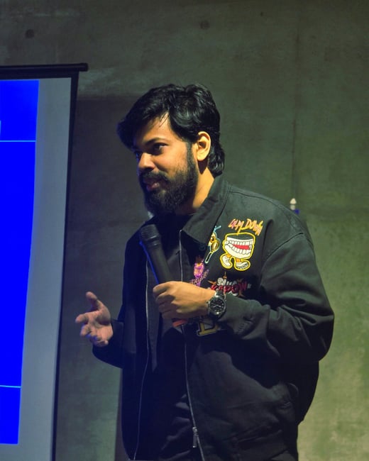 A candid photograph captures a bearded man speaking into a microphone during an indoor presentation, gesturing with his left hand. He is wearing a black jacket with colorful patches and stands in front of a projection screen displaying a solid blue background.