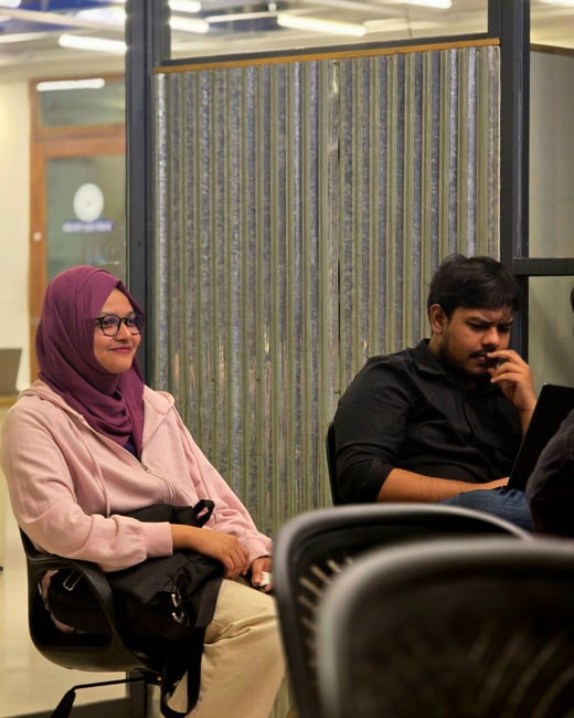 A candid photo captures a young woman wearing a pink hoodie, purple hijab, and glasses smiling while seated, with a man in a black shirt sitting intently beside her in an indoor office or meeting setting.