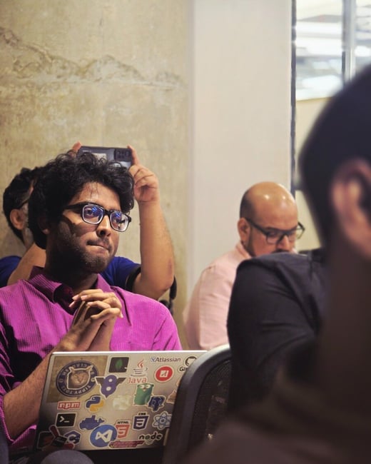 A candid photograph of a young man in a bright magenta shirt and glasses, seated indoors at an event, intently watching a presentation while resting a laptop covered in technical stickers on his lap.