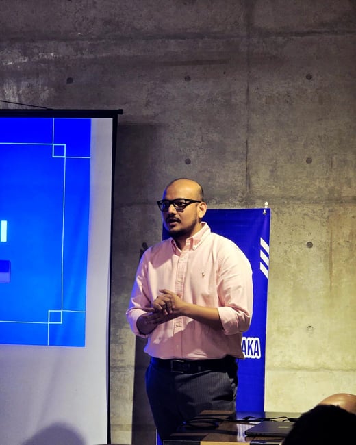 A photograph captures a man wearing glasses and a pink shirt giving a presentation indoors, standing in front of a large blue projection screen and a concrete wall.