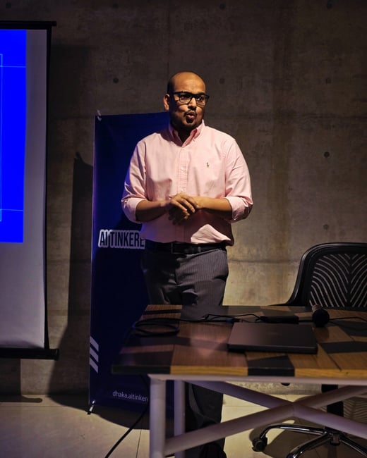 A man wearing a pink button-down shirt and glasses is giving a presentation in a dimly lit indoor setting, standing near a wooden table with a laptop and microphone, and in front of a blue branded banner.