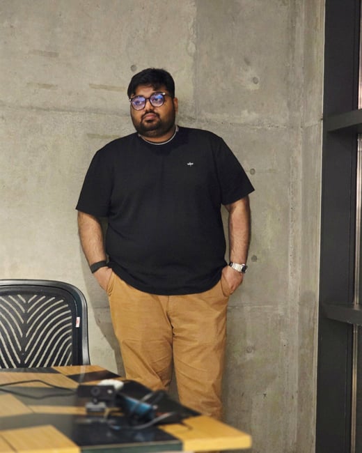 A full-body photograph of a man standing indoors against a raw concrete wall, wearing a black t-shirt and tan trousers with his hands in his pockets.