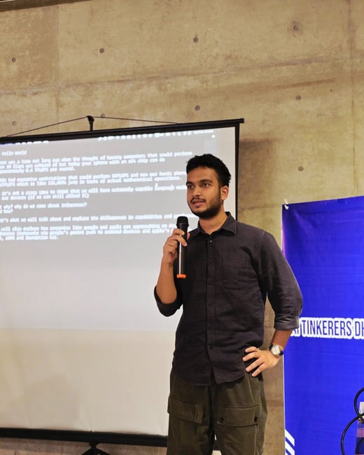 A speaker, a man wearing a dark shirt and cargo pants, stands in front of a large projection screen and a blue banner, holding a microphone while giving a presentation indoors.