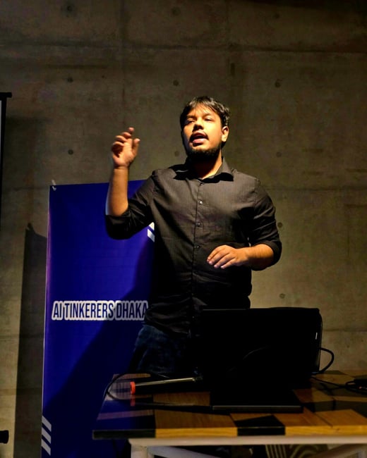 A man wearing a black shirt is giving a presentation, standing behind a wooden table with a laptop, next to a blue banner reading "AI TINKERERS DHAKA." The scene is dimly lit with a concrete wall background.