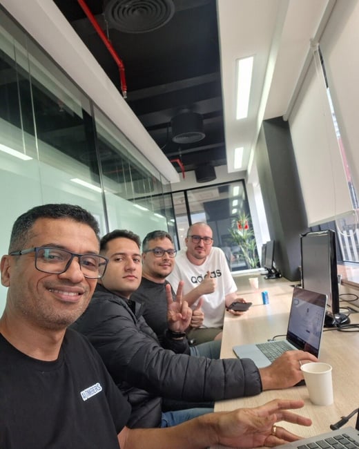 A group of four men posing for a selfie while sitting at a long desk in a modern office environment.