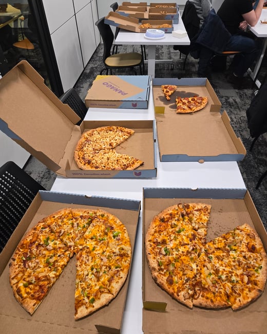 Several open pizza boxes from Panago are spread across white tables in an office-like setting, with some pizzas partially eaten.