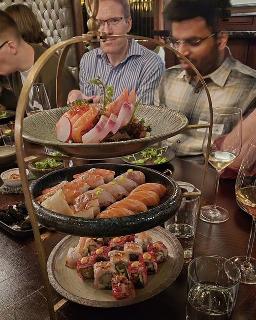 A three-tiered serving stand filled with an assortment of sushi and sashimi sits on a restaurant table surrounded by diners.