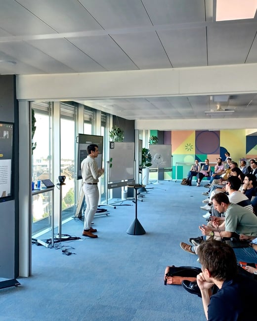 A man is giving a presentation to a seated audience in a modern, well-lit office space featuring large windows and a colorful geometric mural.
