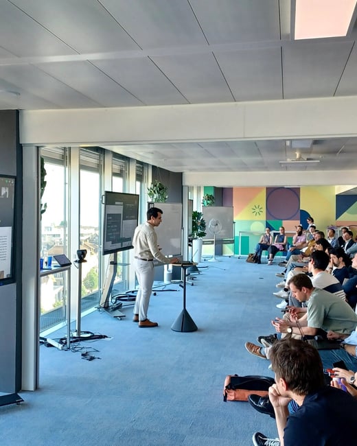 A man is giving a presentation or demonstration to a group of people in a bright, modern office space with large windows.