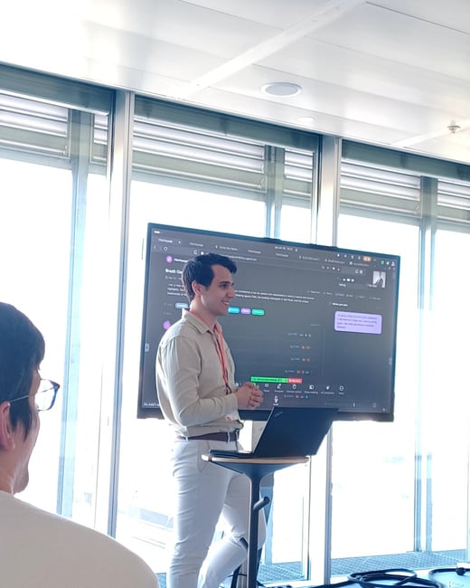 A man in a light-colored shirt is giving a presentation in a bright, modern office, standing next to a laptop on a high stand in front of a large screen showing a digital interface.
