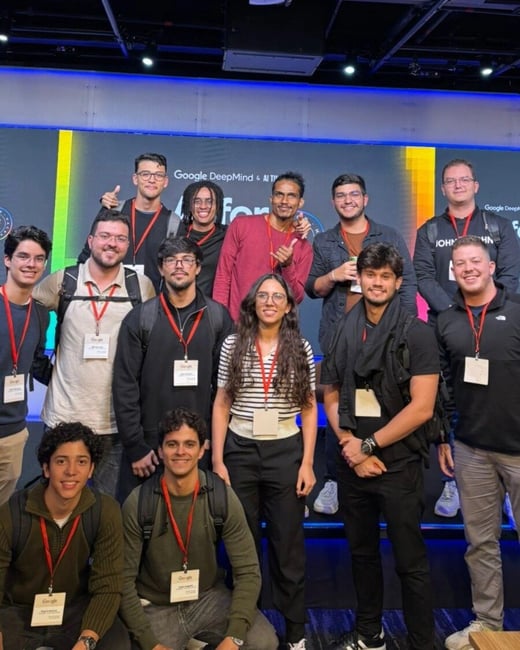 A group of twelve young people posing for a group photo at a Google DeepMind event, standing in front of a large digital screen.