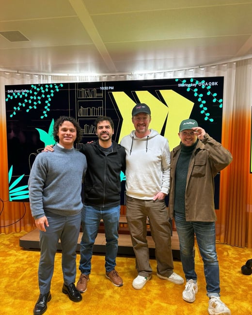 Four men stand shoulder-to-shoulder, posing for a group photo in an indoor setting with a vibrant yellow shag carpet and a large digital screen displaying graphics and text behind them.